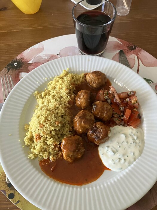 Tallrik med marockanska köttbullar i sås, couscous, yoghurtsås och hackad tomat. Glas med mörkrött dryck i bakgrunden.