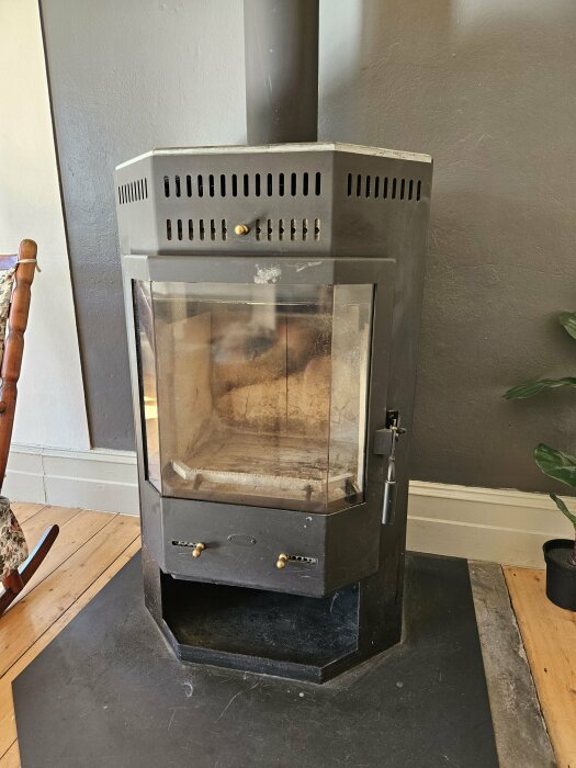 Svart kamin med glaslucka och mässingshandtag placerad i ett hörn av ett rum med trägolv och ljusmålade väggar.