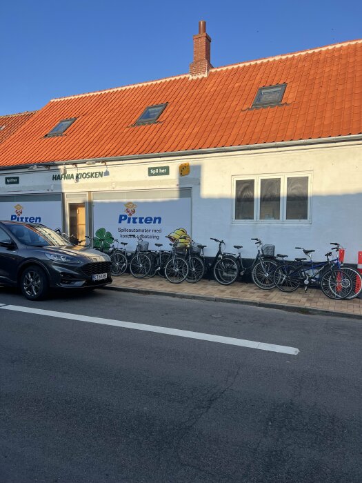 Vit byggnad med orange tegeltak och skyltar med texten "Hanria Kiosken" och "Spil her". Cyklar parkerade framför byggnaden och bil på vägen.