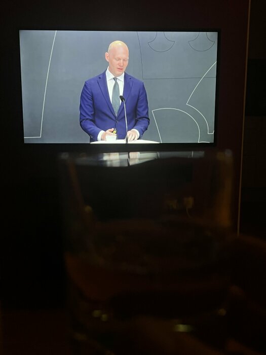 En person i blå kostym talar vid en presskonferens som visas på en tv-skärm. En hand håller ett whiskyglas i förgrunden.