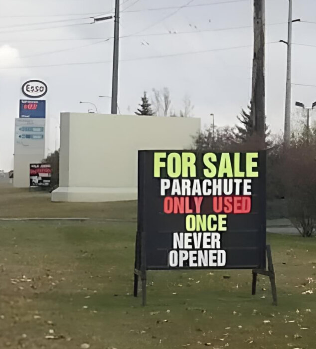 Till salu-skylt för en begagnad fallskärm som aldrig öppnats, med en Esso-bensinstation i bakgrunden.