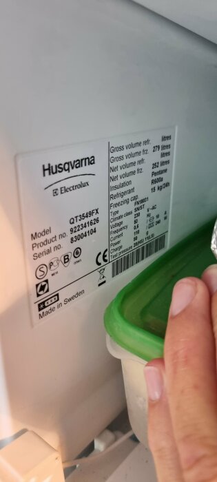 Etikett med teknisk information för en Husqvarna QT3549FX frys, med hand som håller i en plastbricka, isbildning kan anas i bakgrunden.