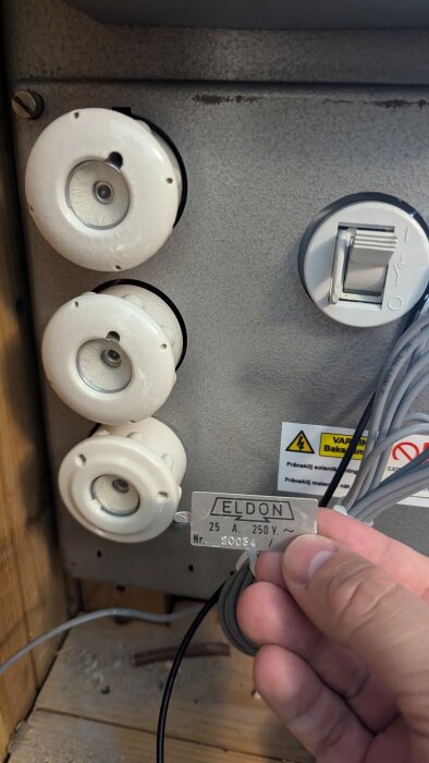 Bild på en mätartavla där en person håller en metallskylt som visar "Eldon 25A 250V", tre säkringar och några exponerade kablar.