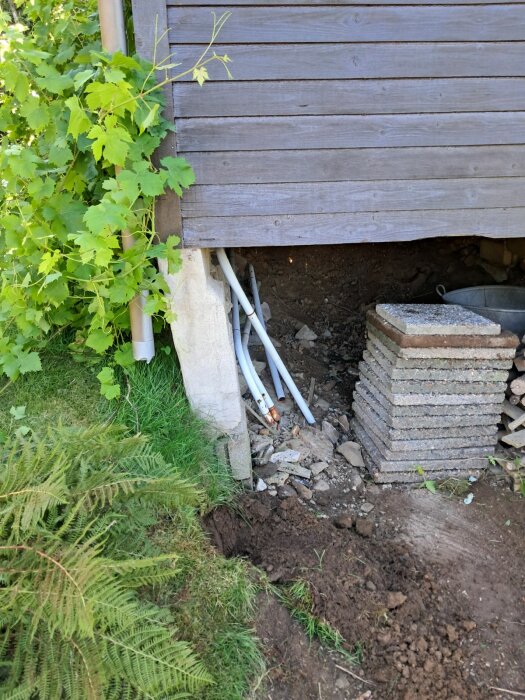 Förvaltning av plint på en friggebod med markvegetation, liggande träpanel och lösa betongplattor bredvid ihoprasad jord.