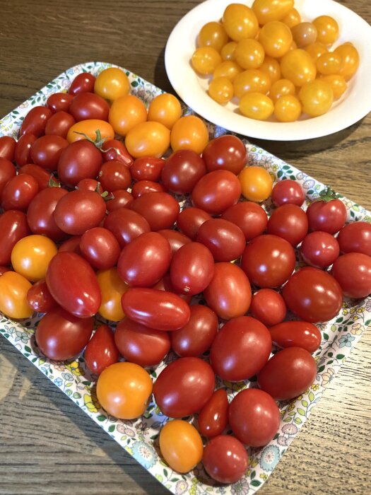 En bricka med röda och orange småtomater samt en vit skål med gula småtomater, placerad på ett träbord.