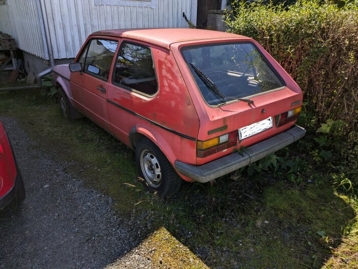 Röd Volkswagen Golf från 1980-talet står oanvänd på en gräsyta intill ett hus, redo för renovering.