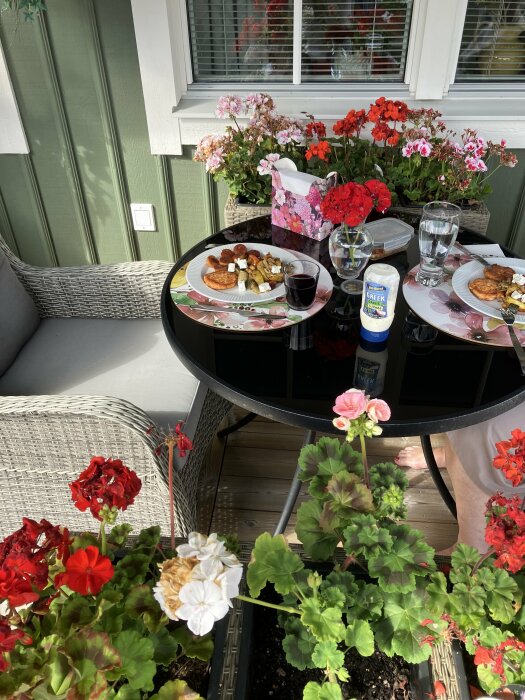 En dukad altan med ett litet bord, två tallrikar med mat, ett glas vin och blommande pelargoner runtomkring.