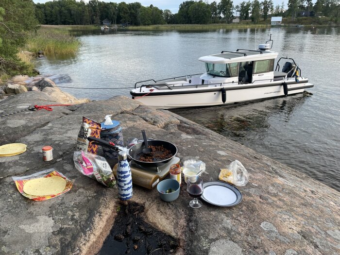 Tacosservering på klippig strand med båt i bakgrunden, matvaror på klipporna inklusive tacobröd, köttfärs, chips och ett glas rödvin, skog i bakgrunden.