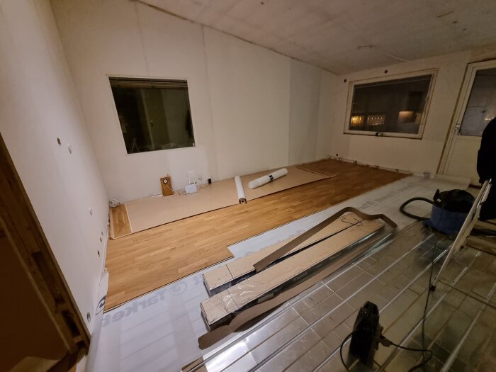 Rum under renovering med vattenburen golvvärme under installation. Träplankor är delvis lagda på golvet, verktyg och material syns i bilden.