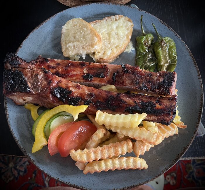 En tallrik med grillade tunna revbensspjäll, pommes frites, stekta padrones, grönsaker och vitlöksbröd med ost.