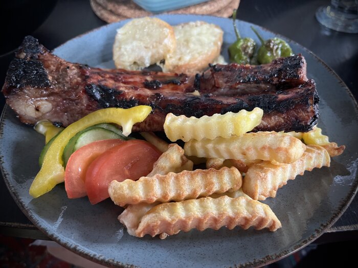 Tallrik med grillade tunna revbensspjäll, pommes frites, stekta padrones, tomat, gurka, paprika och vitlöksbröd toppat med ost.