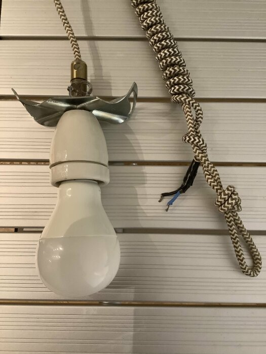 Vit lampa hänger från taket, ansluten via flätad kabel; metalliskt lampholder, ofullständiga trådar syns bredvid, på en vägg med horisontella paneler.