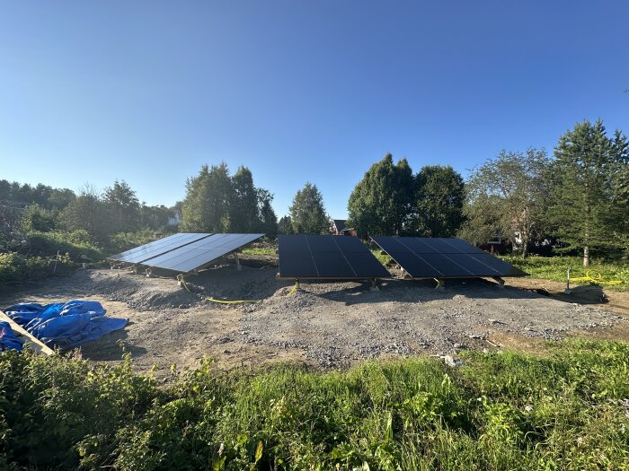 Solpaneler på markställningar på en grusplan med träd och hus i bakgrunden, redo för att kopplas in av en elektriker.