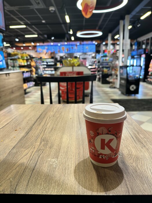 En Circle K pappersmugg med lock står på ett träbord i en butik med drycker och snacks i bakgrunden.