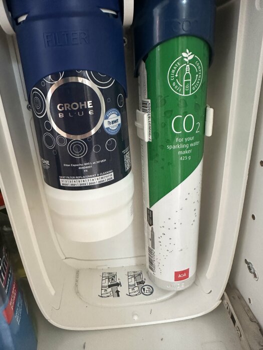 Grohe Blue vattenfilter och AGA CO2-cylinder för kolsyremaskin monterade sida vid sida, fotade i ett skåp.