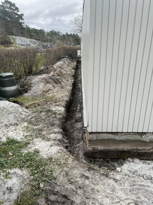 En nygrävd dräneringsdike längs en husvägg med en trädgård i bakgrunden, omgiven av växtlighet och stenig terräng.