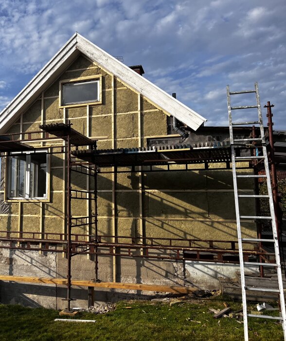 Ett hus under renovering, med isolering och vertikala reglar installerade på ytterväggen. Ställningar och stegar finns runt huset. Klar himmel med moln.