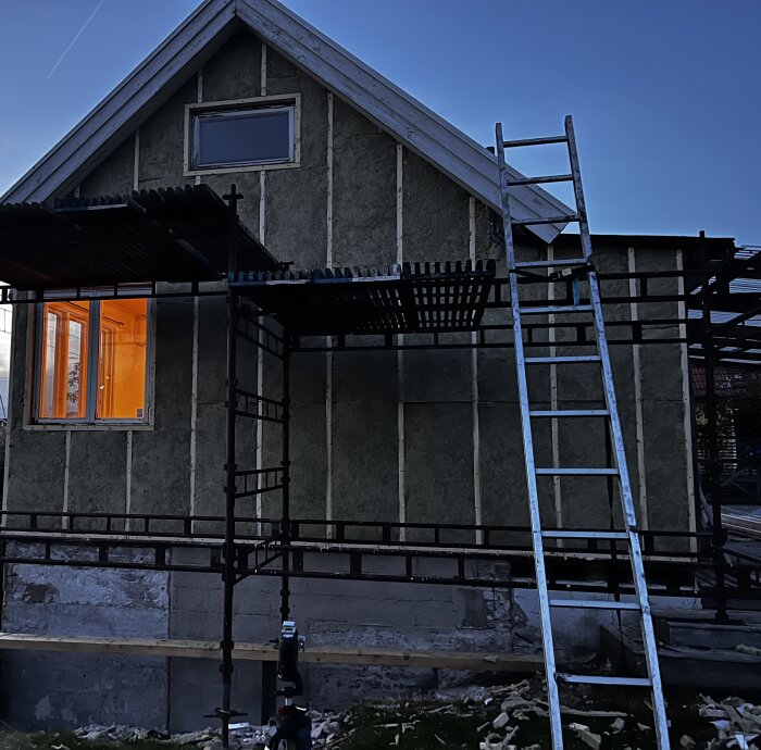 Ett hus under renovering med ställningar runt fasaden. Isoleringsmaterial har installerats och en byggstege står lutad mot huset. Ett fönster lyser med varmt sken.