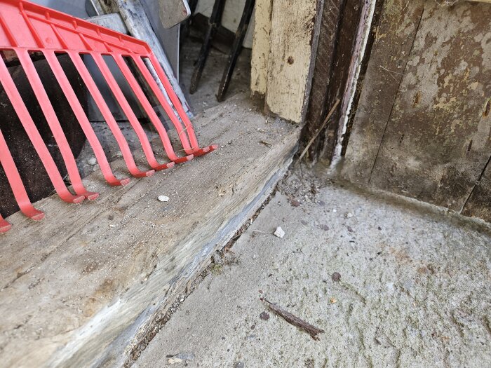 Ett hörn av en gammal byggnad med sliten trästock, delvis nedsjunken i marken. En röd plastkratta vilar på stocken, och marken är grusig och ojämn.