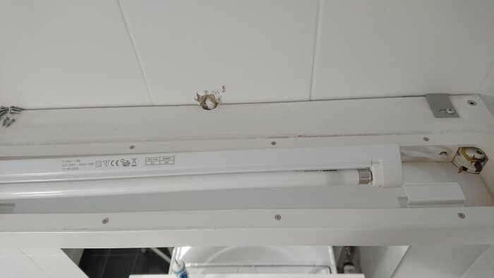 Demonterad belysningsarmatur i ett spegelskåp, med synliga kablar och en exponerad vägg bakom; skruvar ligger på sidan av armaturen.