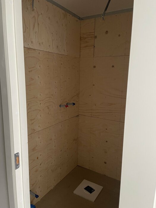 Ett rum under konstruktion med plywoodväggar och förberedelser för att installera vattenrör och golvbrunn.