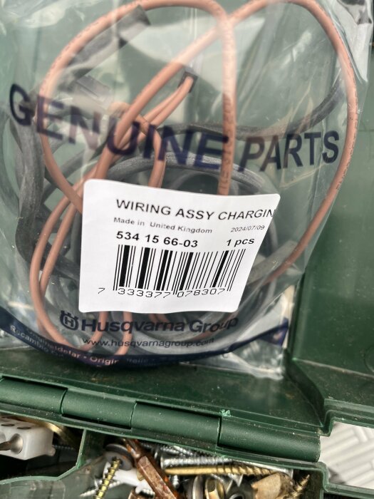 Husqvarna laddningskabelsats, artikelnummer 534 15 66-03, i en plastpåse märkt "Genuine Parts", placerad i en grön verktygslåda med skruvar och hårdvara.