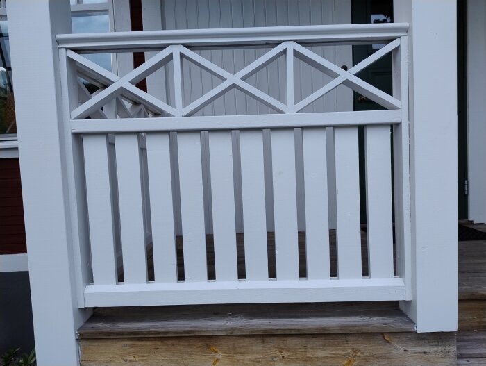 Ett vitt balkongräcke med vertikala ribbor och korslagda ribbor upptill, monterat på en veranda med trätrappa.