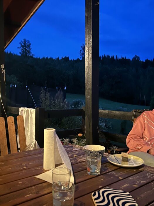 Bilden föreställer en uteplats på kvällen med ett bord fullt av glas och tallrikar, en stolpe i mitten och en mörk natthimmel i bakgrunden.