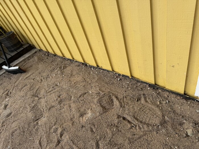 Gult träfasadhus med en smutsig mark bestående av sand och sten, ingen betongram vid husväggen, sopborste synlig till vänster.