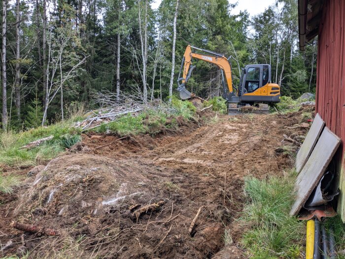 En grävmaskin gräver på en tomt bredvid en röd lada och avslöjar en stor sten eller berg. Området är omgivet av skog.