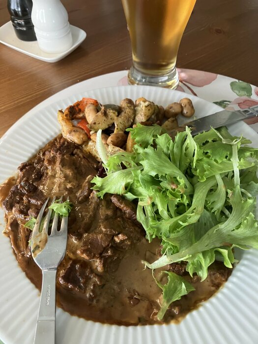 En tallrik med långkokt lövbiff i sås, grönsaker och sallad, med ett glas öl i bakgrunden.