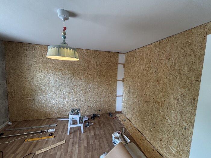 Ett rum under renovering med osb-skivor på väggarna, en hängande lampa i taket och verktyg samt en pall på golvet.