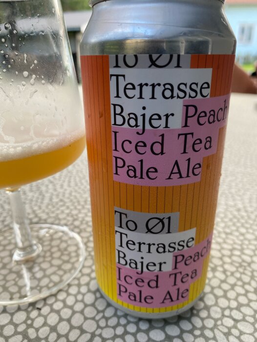 En ölburk med etiketttexten "To Øl Terrasse Bajer Peach Iced Tea Pale Ale" och ett glas halvfullt med öl på ett bord utomhus.