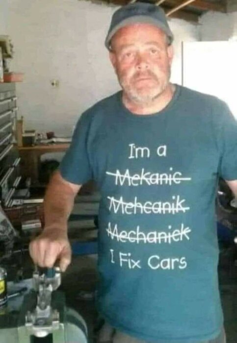 En man med en keps står i ett garage och bär en t-shirt med texten "I'm a Mekanic Mehecanik Mechanick I Fix Cars.