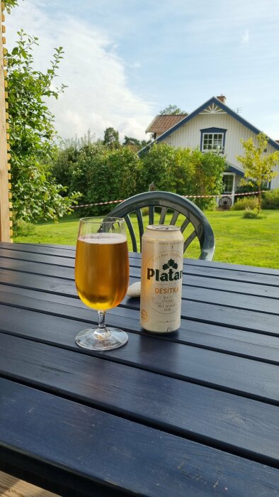 En öppnad burk Platan Desítka öl och ett glas med öl på ett blått träbord utomhus, med en vit träbyggnad och grön trädgård i bakgrunden.