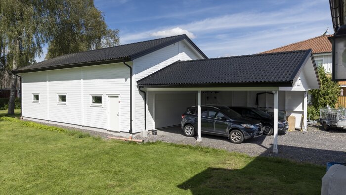 Vittrmålat hus med svart tak och vindskivor, bredvid en carport med två parkerade bilar, omgivna av en gräsmatta under en klarblå himmel.