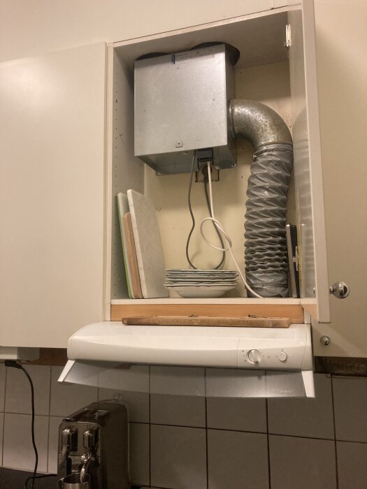 Köksskåp med borttagen lucka som visar en äldre fläktinstallation med metallbox och ventilationsrör, skärbrädor och tallrikar står ovanför en vit spiskåpa.