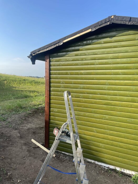 Stege lutad mot en grön byggnad med nyinstallerade knutbrädor och vindskivor på ena sidan. Grönskande fält och klar himmel i bakgrunden.