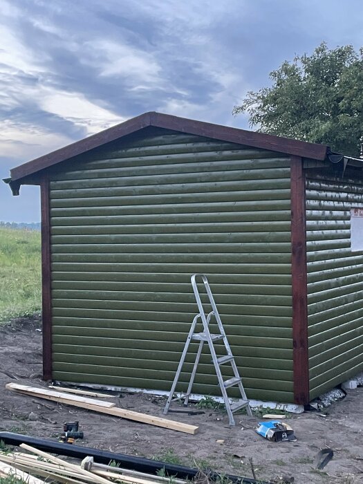 En sida av ett grönt byggnad med nyligen installerade knutbrädor och vindskivor, stege och byggverktyg på marken runt omkring.