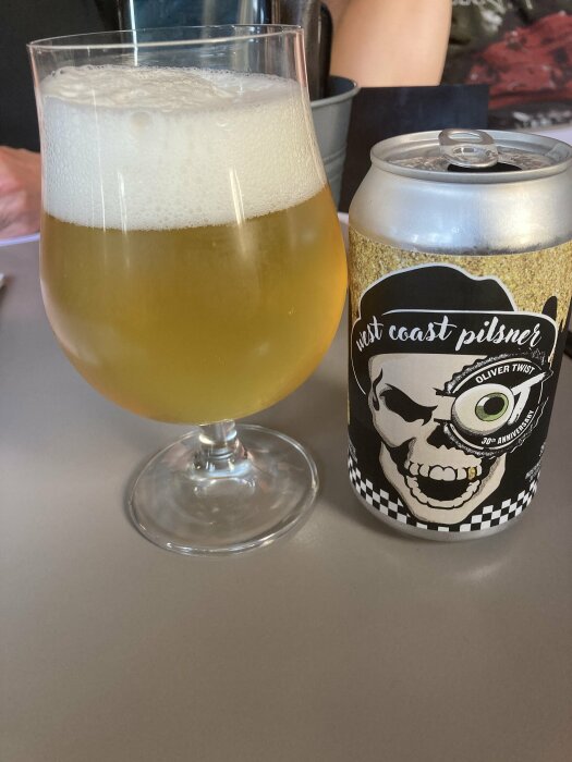 Ett glas med gyllengul öl med skum och en ölburk märkt "West Coast Pilsner" med ett dödskallemotiv på ett bord.