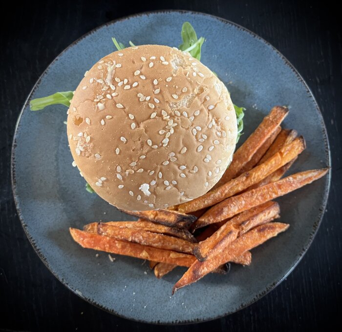 Hamburgare med sesamfröbröd och grönsaker serverad med sötpotatispommes på en grå tallrik.