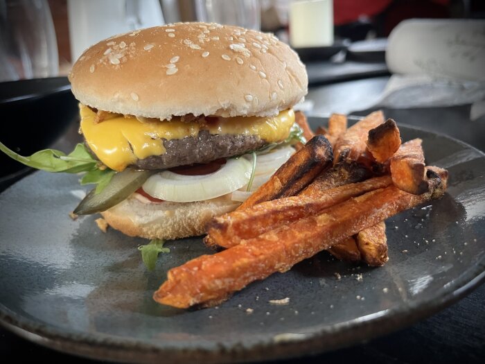 En tallrik med en hamburgare med sallad, lök, ost och dressing, och sötpotatispommes frites bredvid, serverad på ett bord.