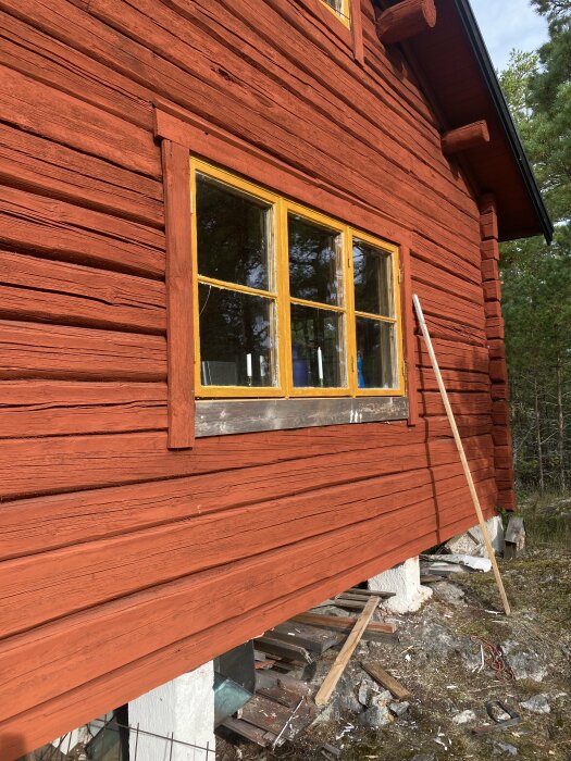 Rött trähus med ett fönster där fodret är borttaget, avslöjande en skada under fönstret. Några verktyg och träbitar på marken under.