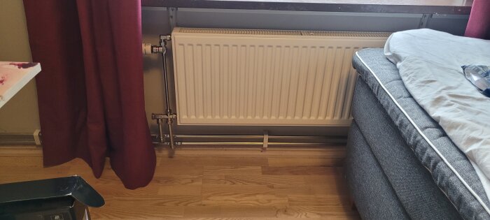 Nyinstallerat vattenelement kopplat till bergvärme bredvid en säng i ett sovrum, med röda gardiner och trägolv.