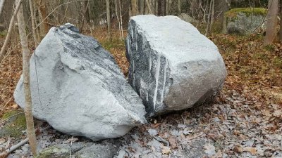 Två stora spräckta stenblock ligger på marken omgivna av växtlighet och höstlöv.