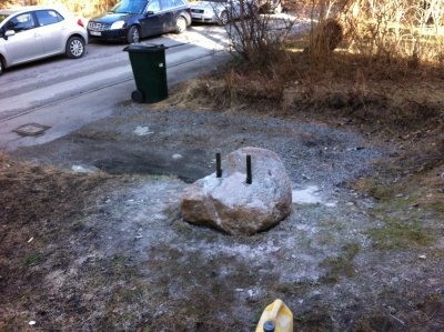 Stenblock med två metallstänger insatta, troligtvis för att klyvas. Området runt stenen är grusigt. I bakgrunden syns bilar och en soptunna vid vägkanten.