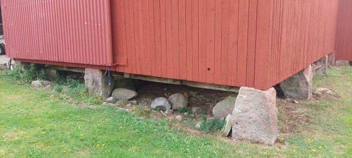 Röd byggnad på ojämna grundstenar, bärande på stora stenar. Gräns mellan trävägg och gräs med synlig nivåskillnad och vegetation runt stenar.