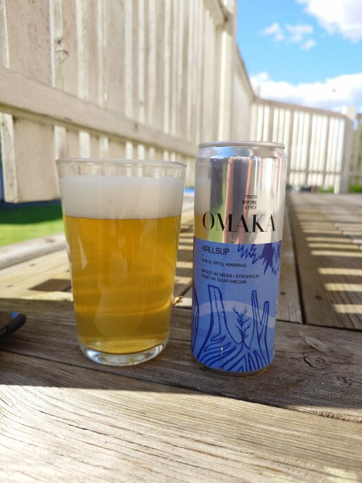 Ett glas öl och en burk märkt "OMAKA Kallsup" på ett trädäck med en vit staketbakgrund under en solig himmel.