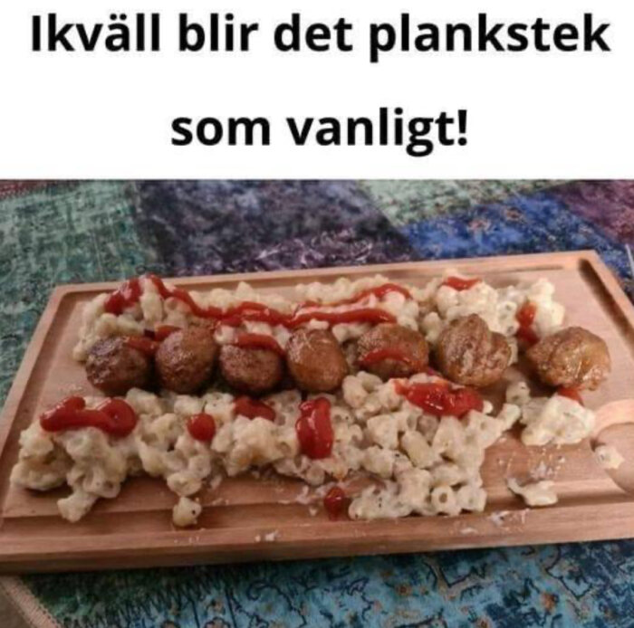 Planka med köttbullar och gratinerad pasta, toppad med ketchup, ligger på ett träplank. Texten ovanför säger "Ikväll blir det plankstek som vanligt!