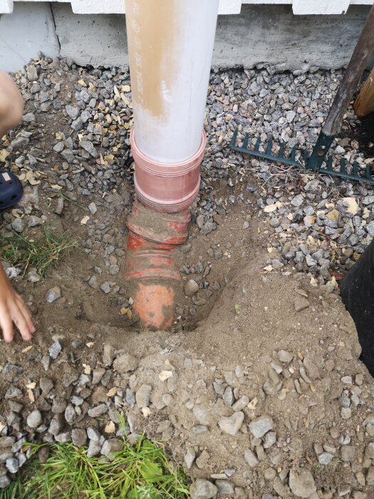 Ett grävt hål vid en husvägg visar en vit och orange dräneringsrör som svänger 90 grader under marknivån. Runt röret syns jord och småstenar.